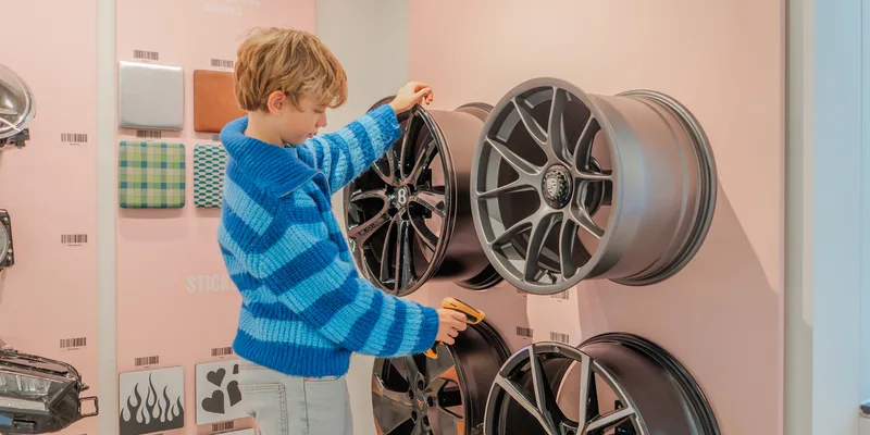 A child is standing in front of different wheel caps. It is holding a hand scanner and is scanning a bar code underneath one of the wheel caps.